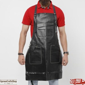Könnte beinhalten: Schwarze Lederschürze mit weißen Nähten, zwei großen Vordertaschen und einer Brusttasche. Die Schürze hat verstellbare Träger und einen Schnallenverschluss. Die Schürze ist für den Einsatz in einer Werkstatt oder Küche konzipiert. Auf der Schürze steht "ApronsCraftsShop".