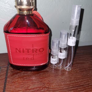 Puede incluir: Un frasco de perfume rojo con tapa dorada y el texto "NITRO pour homme red" en una etiqueta de cuero roja. Tres frascos rociadores transparentes, etiquetados 3 ml, 5 ml y 10 ml, están junto al frasco de perfume. El frasco de perfume tiene una correa de cuero.