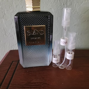 Puede incluir: Un frasco de perfume con una tapa dorada y un diseño degradado azul oscuro y plateado. El frasco tiene las palabras "ISLAND DREAMS KHADIJAH" en una etiqueta dorada. Varias botellas de spray pequeñas y transparentes están junto al frasco de perfume, etiquetadas 3ML, 5ML y 10ML.
