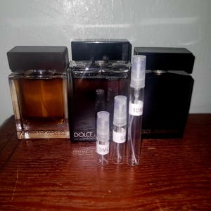 Peut inclure: Trois flacons de parfum rectangulaires en verre avec des bouchons noirs et trois petits vaporisateurs cylindriques transparents étiquetés 3ML, 5ML et 10ML. Les flacons portent la marque Dolce & Gabbana, avec un liquide ambré et deux liquides foncés.