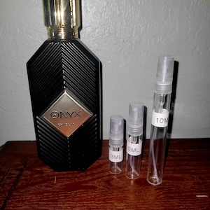 May include: A black perfume bottle with a gold cap and a diamond-shaped label reading ONYX KHADLAJ, accompanied by three clear glass spray vials labeled 3ML, 5ML, and 10ML, arranged in a row next to the larger bottle on a dark wooden surface.