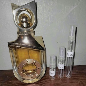 Könnte beinhalten: Eine goldfarbene Parfümflasche mit einem einzigartigen geometrischen Design, beschriftet mit "ZENITH", wird neben drei kleinen, transparenten Sprühflaschen mit den Aufschriften 3ML, 5ML und 10ML gezeigt. Die Parfümflasche hat einen dekorativen Aufsatz.
