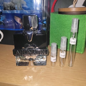 Puede incluir: Un frasco de perfume de vidrio negro con tapa plateada y una banda plateada con el texto AROMATIX FRENCH AVENUE, acompañado de tres viales de spray de vidrio transparente etiquetados como 3ML, 5ML y 10ML, ordenados por tamaño.