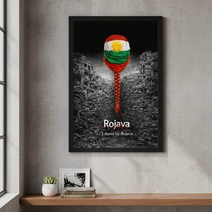 May include: Framed art print with a flag-themed design on a head, with a braid, set against a backdrop of a destroyed city. The flag incorporates red, green, yellow, and white. Text reads "Rojava I stand by Rojava."