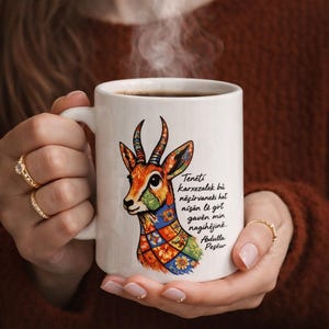 May include: White ceramic coffee mug with a colourful gazelle illustration and text. The gazelle has a patterned neck and head with orange, blue, green, and yellow accents. The mug is filled with dark coffee, and steam rises from the top.