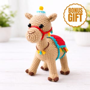 Könnte beinhalten: Ein gehäkeltes Kamelspielzeug in Beige mit roter Trense, rotem und blauem Sattel und einem kleinen Hut. Das Kamel hat schwarze Augen und eine dekorative Halskette. Das Bild enthält einen "BONUS GIFT"-Aufkleber.