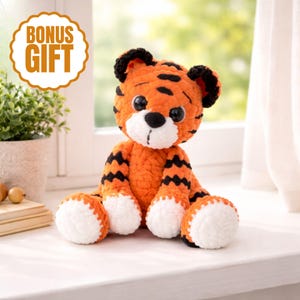 May include: A crocheted tiger toy in orange and black, with white details. The tiger features large black eyes, a white snout, and black stripes. The image includes a bonus gift label.