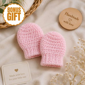 May include: Two light pink, hand-crocheted baby mittens. The mittens are displayed on a soft, cream-colored surface, with a wooden disc that reads "Handmade with love." A card reads "Made with love for your little one." A bonus gift sticker is visible.