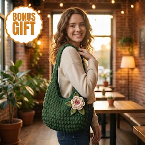 May include: A dark green crochet shoulder bag with a large flower applique. The flower has pink and green petals. The bag is made of thick yarn and has a rounded shape. The image also includes the text "BONUS GIFT" in a gold circle.