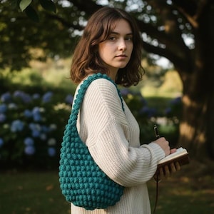 May include: A teal green, crocheted shoulder bag with a textured bobble stitch design. The bag is worn over the shoulder. The person is wearing a cream-coloured sweater and holding a notebook and pen.