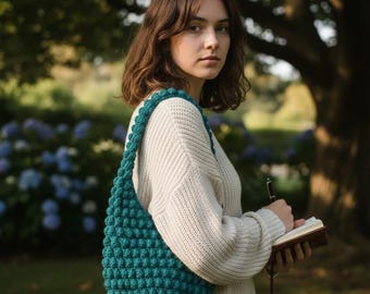 May include: A teal green, crocheted shoulder bag with a textured bobble stitch design. The bag is worn over the shoulder. The person is wearing a cream-coloured sweater and holding a notebook and pen.