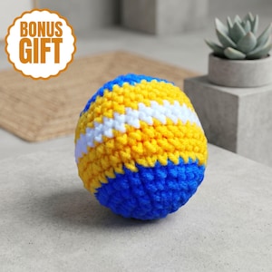 May include: A spherical crochet ball featuring horizontal stripes of bright blue, vibrant yellow, and white yarn. The textured surface is created with a consistent knit pattern, showcasing a rounded shape with alternating colour bands throughout.