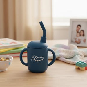 May include: A navy blue sippy cup with a lid and a flexible straw. The cup has two handles and a white cartoon crocodile design. The cup is sitting on a wooden table next to a bowl of cereal, a stack of books, and a framed photo.
