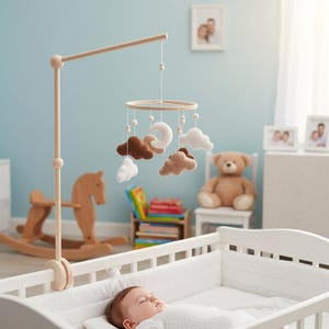 May include: A wooden baby mobile with felt clouds, moons, and a wooden ring hangs above a white crib. The mobile is supported by a wooden stand. A baby sleeps peacefully in the crib. The room has a light blue wall.