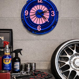 May include: A blue and pink clock made from a car clutch assembly. The clock has pink numerals and hands, with a blue outer ring. The clock is mounted on a grey brick wall. Automotive theme.