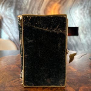 Puede incluir: Un pequeño libro antiguo rectangular con una cubierta de terciopelo negro y un borde dorado. El libro está cerrado y tiene un pequeño cierre en el lateral. Los bordes del libro muestran signos de desgaste.