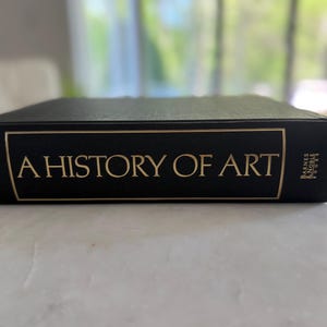 May include: A History of Art book with a black cover and gold lettering. The title is prominently displayed, with the author's name and publisher's logo visible. The book is lying flat on a light-colored surface.