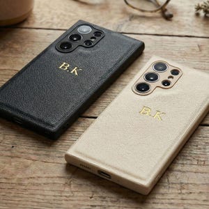 Puede incluir: Dos fundas de teléfono de cuero, una negra y otra crema, se muestran sobre una superficie de madera. Cada funda presenta un monograma dorado "B.K." y un recorte para el módulo de la cámara. Las fundas tienen un acabado texturizado.