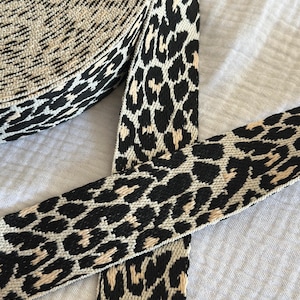 Puede incluir: Un rollo de cinta tejida con un patrón repetitivo de leopardo en negro y beige. La tira de tela tiene una superficie texturizada con un ancho constante y un tejido denso y duradero, presentada como una bobina enrollada con un extremo.