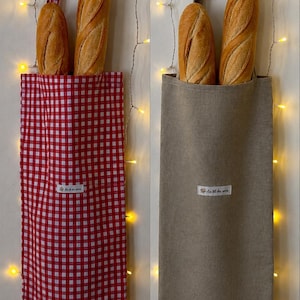May include: Two tall fabric baguette bags, one in a red and white gingham check pattern and the other in a solid beige color. Each bag features a long shoulder strap and a small white rectangular label with the text Au fil des rêves.