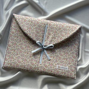 Puede incluir: Un estuche tipo sobre con estampado floral y cierre de lazo de cuadros vichy azul claro. El estuche presenta un patrón de pequeñas flores rosas sobre un fondo crema. Una pequeña etiqueta con texto es visible en la esquina inferior derecha.