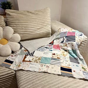 Può includere: Una coperta patchwork per bambini con vari modelli di tessuto, tra cui strisce, stampe di animali e temi nautici. La coperta è posizionata su un divano di velluto a coste beige, insieme a un cuscino a forma di margherita e un cuscino rettangolare.