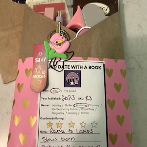 Puede incluir: Un paquete de papel marrón con una banda rosa con estampado de corazones, que contiene artículos relacionados con libros. Una etiqueta indica el género del libro como romance, publicado en 2023. Un llavero de flor rosa y pegatinas son visibles.