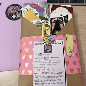 May include: A brown paper package wrapped with a pink band featuring gold hearts. The package includes a label titled BLIND DATE WITH A BOOK, listing 2021 as the year published, SKU M14, and Mystery/Thriller genre. It includes stickers, a pen, and a book charm.