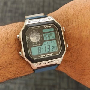 May include: A Casio digital watch with a blue strap is shown. The watch face is black with a digital display showing the time, date, and other functions. The watch has the text "WORLD TIME", "CASIO", "10 YEAR BATTERY", and "ILLUMINATOR".