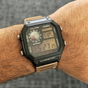 May include: Black and gold Casio digital watch with a rectangular face. The watch displays the time, date, and world time functions. The watch band is gold-colored. The watch has the text "CASIO", "10 YEAR BATTERY", and "ILLUMINATOR".