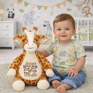 May include: A plush giraffe toy with brown and white spots, personalized with the name "James" and birth details. The giraffe is next to a baby wearing a green shirt with animal graphics and blue jeans. The background features a nursery theme.