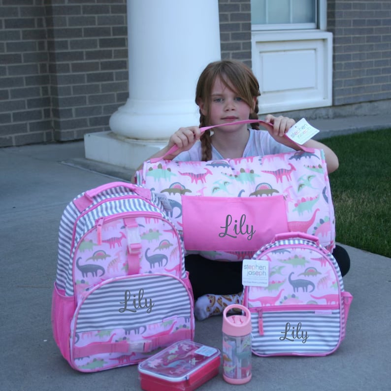 May include: A pink and gray backpack with a dinosaur pattern and the name "Lily" embroidered on it. There is also a matching lunch bag, water bottle, and a pink and white duffel bag with the name "Lily" embroidered on it.