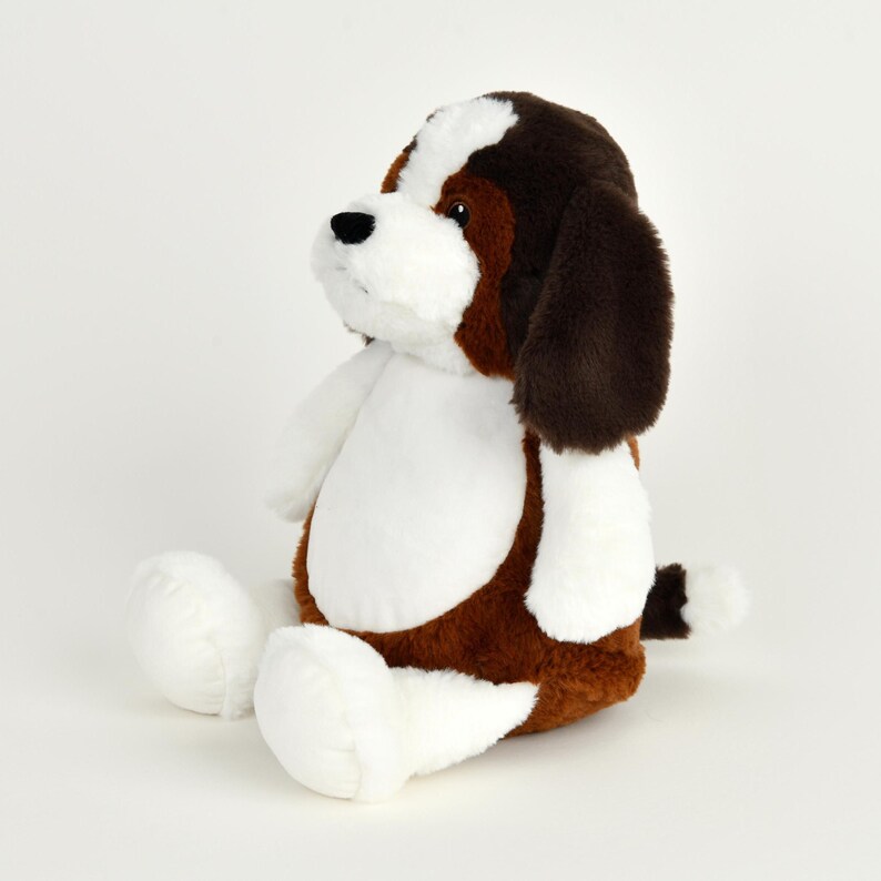 May include: A plush stuffed dog toy in a sitting position. The toy features a brown and white color scheme, with brown ears and patches, and white accents on the chest, paws, and tail. The dog has a black nose and is set against a white background.