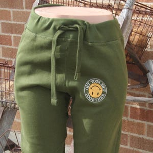 May include: Olive green sweatpants with a drawstring waist and a smiley face graphic that says "GET YOUR SHIT TOGETHER". The pants are displayed on a mannequin and have a relaxed fit.