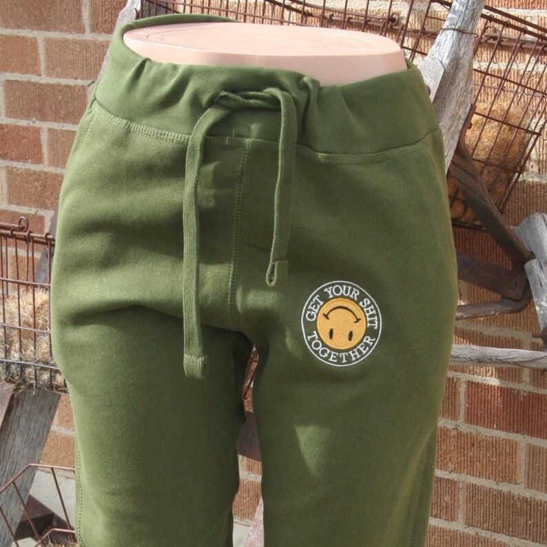 May include: Olive green sweatpants with a drawstring waist and a smiley face graphic that says "GET YOUR SHIT TOGETHER". The pants are displayed on a mannequin and have a relaxed fit.