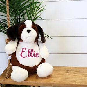 May include: A plush St. Bernard puppy dog with brown, white, and black fur. The dog has the name "Ellie" embroidered in pink on its chest. The stuffed animal is sitting on a wooden swing.