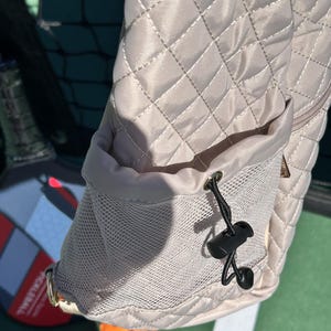 K&ouml;nnte beinhalten: Eine beige gesteppte Tasche mit einem Netzfach und einem Kordelzug. Der Kordelzug hat einen schwarzen Plastikverschluss.