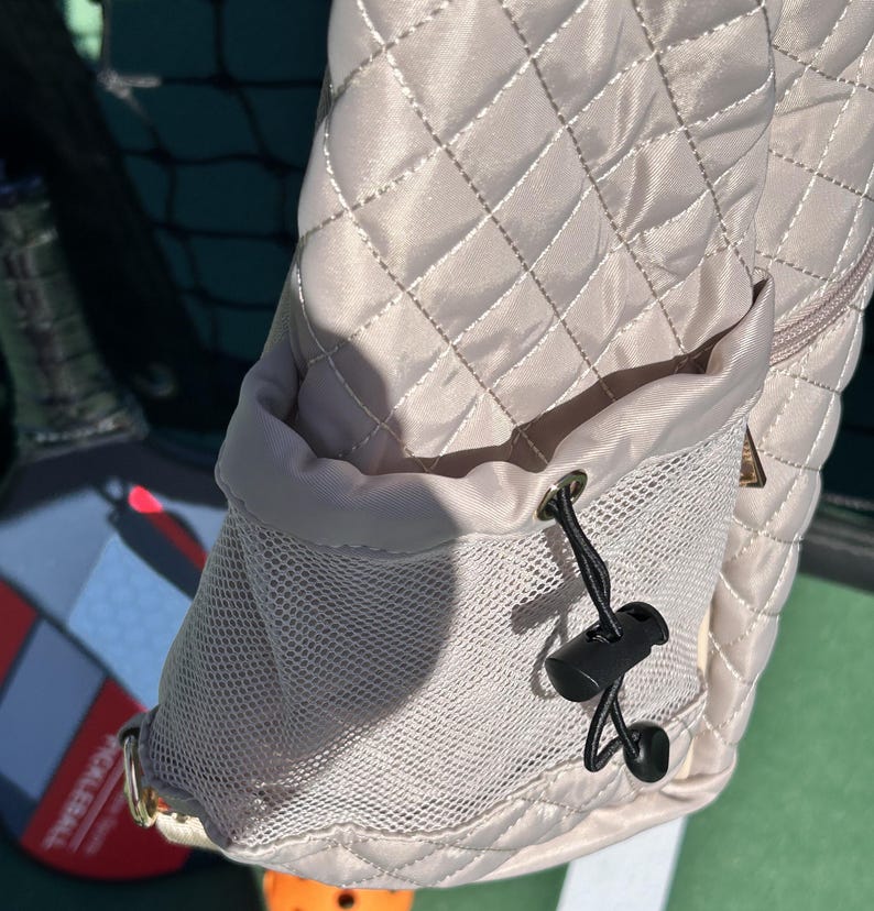K&ouml;nnte beinhalten: Eine beige gesteppte Tasche mit einem Netzfach und einem Kordelzug. Der Kordelzug hat einen schwarzen Plastikverschluss.