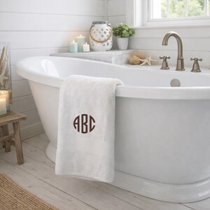 May include: A white bath towel featuring a dark brown circular monogram with the letters ABC embroidered in the center. The towel is draped over the edge of a white freestanding bathtub in a bathroom setting with candles and a small plant.