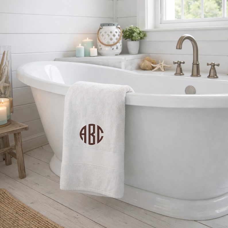 May include: A white bath towel featuring a dark brown circular monogram with the letters ABC embroidered in the center. The towel is draped over the edge of a white freestanding bathtub in a bathroom setting with candles and a small plant.