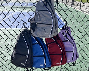 May include: Five different colored sling backpacks hanging on a chain link fence. The backpacks are all the same style and have a white zipper and white trim. The backpacks are gray, black, blue, red, and purple. The gray backpack has the initials "JLA" embroidered on it.