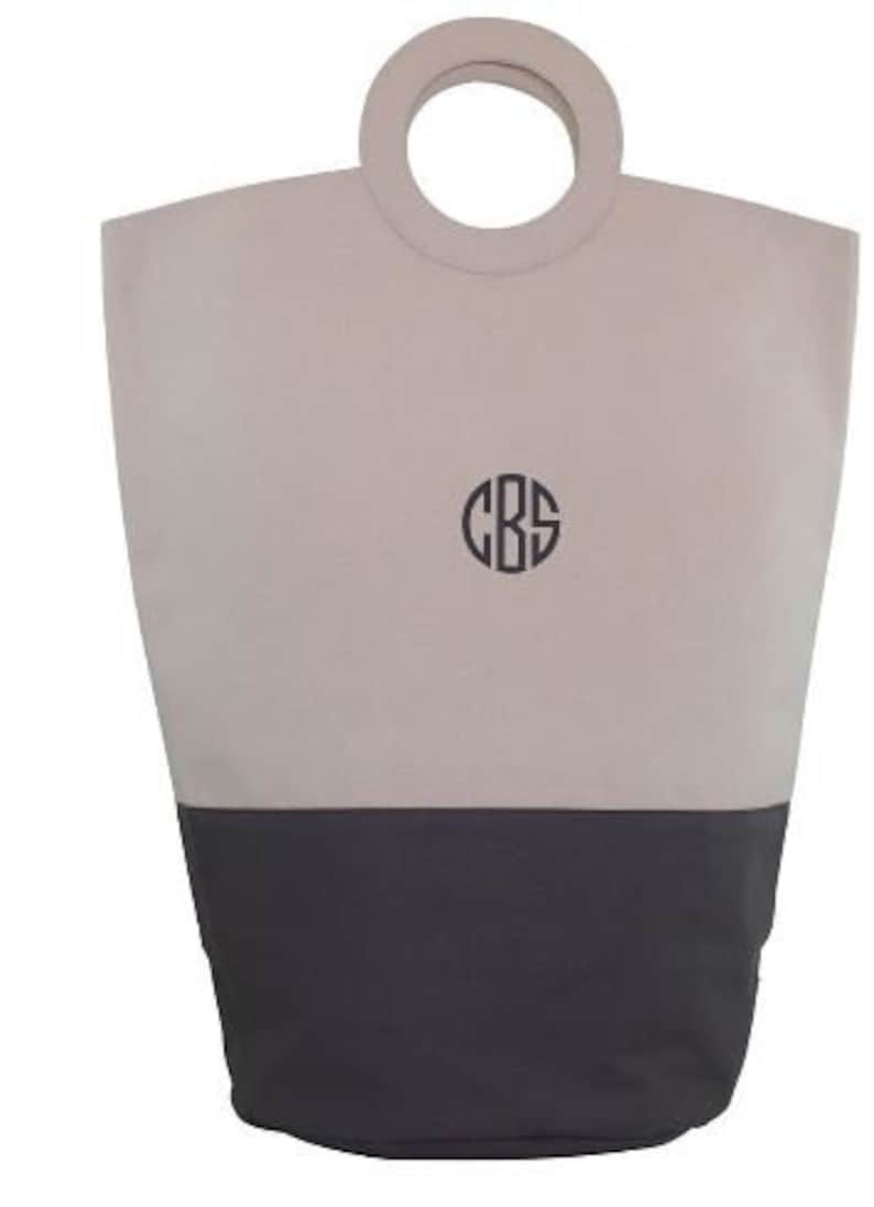 May include: A beige and dark gray canvas tote bag with a large, round, wooden handle. The bag has a monogram of the letters "CBS" in a circle on the front.