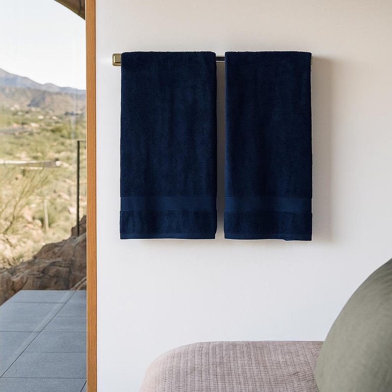 May include: Two rectangular navy blue bath towels hanging side by side on a horizontal metal towel bar. Each towel features a smooth, solid-colored fabric texture with a subtle horizontal band detail near the bottom edge of the textile.