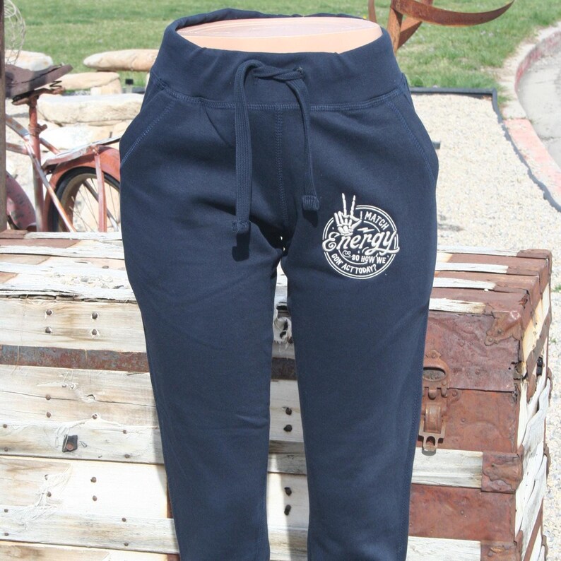 May include: Navy blue sweatpants with a drawstring waist and pockets. A white graphic on the left leg reads "Match Energy" with a hand gesture and the text "Go Do BSW We Act Today". The pants are made of a soft material.