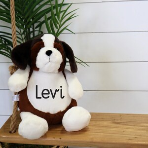 May include: A plush St. Bernard puppy dog with brown, white, and black fur. The dog has the name "Levi" printed on its chest. The stuffed animal is sitting on a wooden swing.