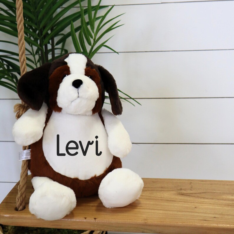 May include: A plush St. Bernard puppy dog with brown, white, and black fur. The dog has the name "Levi" printed on its chest. The stuffed animal is sitting on a wooden swing.