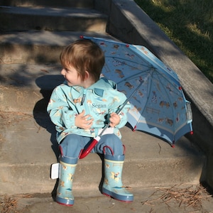 May include: A young child wearing a blue raincoat with a horse and rider design and the name "Segan" embroidered on the front. The child is holding a blue and white umbrella with a horse and rider design. The child is wearing blue rain boots with a horse and rider design.