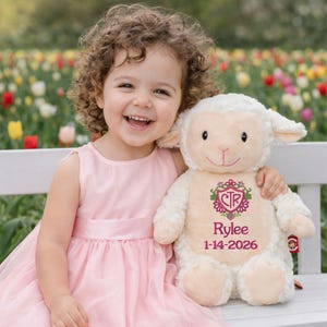 May include: A plush cream-colored lamb stuffed animal featuring embroidered details on its chest, including a pink floral crest with the letters CTR, the name Rylee, and the date 1-14-2026 in pink thread, with soft fabric and floppy ears.