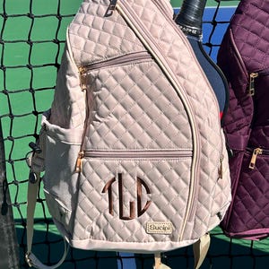K&ouml;nnte beinhalten: Eine gesteppte beige Tennistasche mit einem Rei&szlig;verschlussfach und einem Monogramm "TLD" in braunem Garn. Die Tasche hat einen goldenen Rei&szlig;verschluss und der Markenname "Sucipi" ist auf dem Boden aufgedruckt.