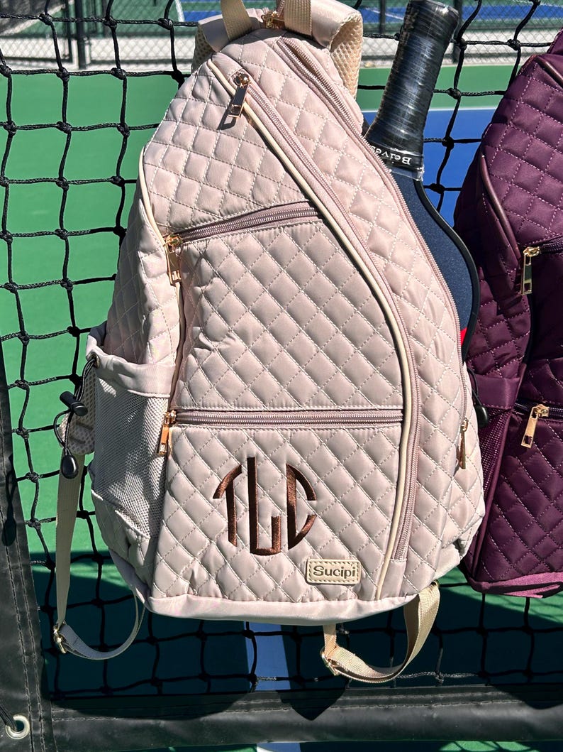 K&ouml;nnte beinhalten: Eine gesteppte beige Tennistasche mit einem Rei&szlig;verschlussfach und einem Monogramm "TLD" in braunem Garn. Die Tasche hat einen goldenen Rei&szlig;verschluss und der Markenname "Sucipi" ist auf dem Boden aufgedruckt.